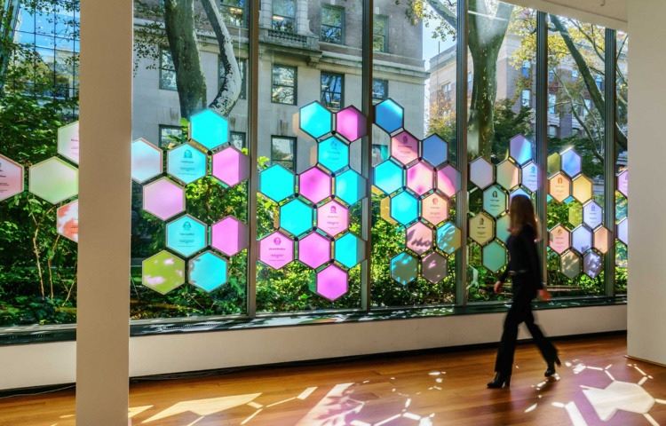 Rockefeller University art instillation highlighting Pearl Meister Greengard Prize winners. It features colorful translucent hexagons hanging in front of a window that display past winners. 