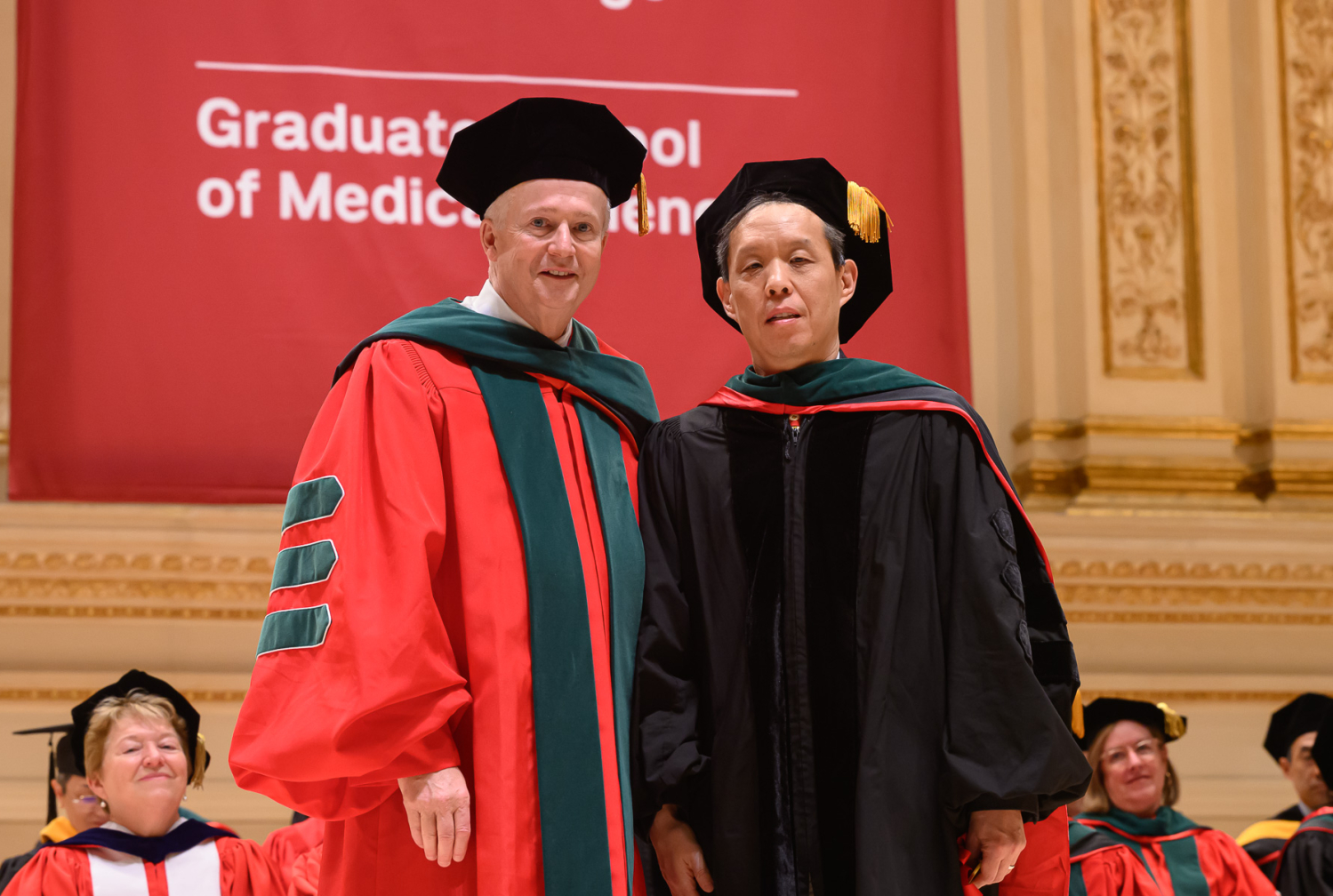 Two men wearing graduation regalia posing during commencement exercises.