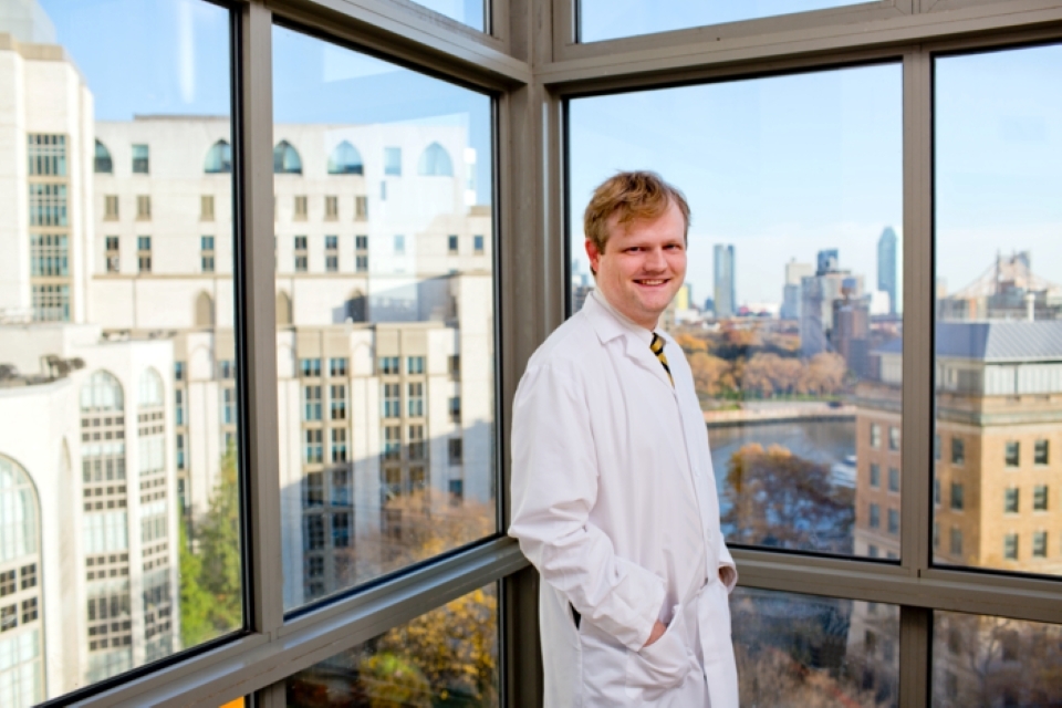 A physician-scientist wearing a white coat stands in front of a window overlooking buildings.