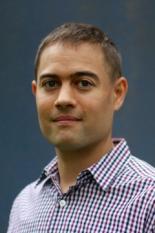 a fair-skinned man wearing a plaid shirt posing for a portrait in front of a blue background.