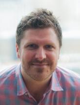 a fair-skinned man seated wearing a plaid shirt posing for a portrait.